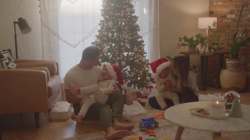 A family is gathered around a litup christmas tree in a living room with a father and mother sitting on the floor with their two small children all four are wearing red and white santa hats the parents are playing with the children who are dressed in