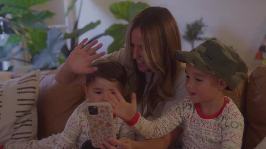 A young mother and her sons wave goodbye while on a video call with a phone in a living room during daytime