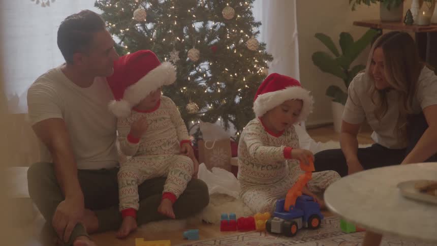 Parents watch smiling as children play with blocks in front of the christmas tree