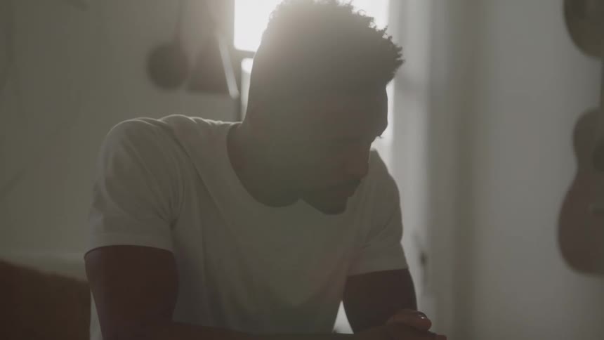 A young man wearing a white tshirt sits indoors with a guitar hanging on the nearby wall at midday