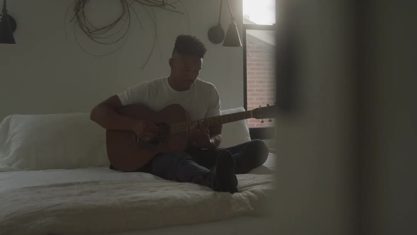 Man sitting on the bed playing guitar in a room lit by the window