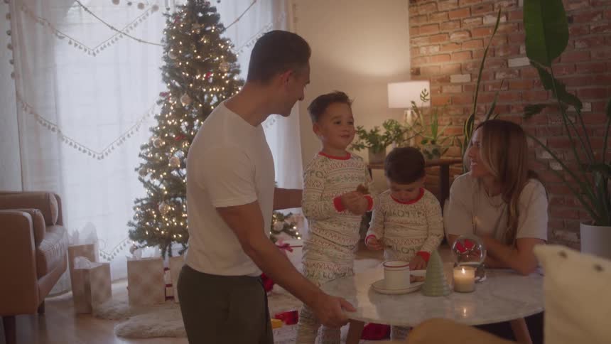 A family gathered at christmas in a living room decorated for the holiday the father mother and two children at the table with the boys eating cookies