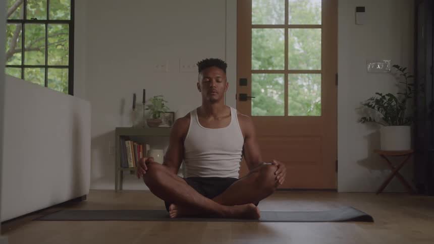 A young black man wearing a white singlet sits indoors meditating with his eyes closed