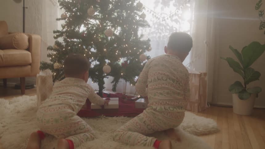 Two children in pajamas look at gifts under a christmas tree
