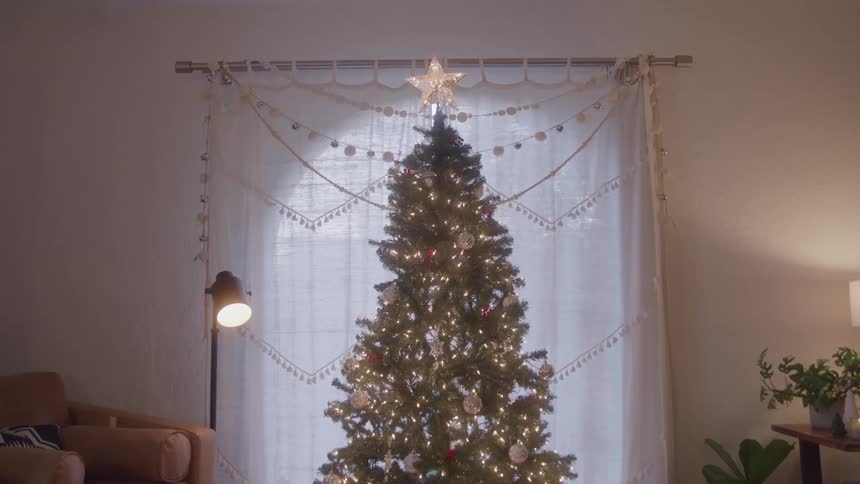 A large christmas tree in a living room in front of a window covered by a white curtain