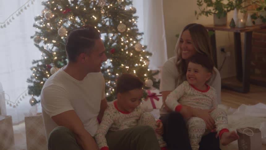 A family gathered in the living room near a christmas tree with one boy on his mothers lap and another boy near his father all talking and smiling