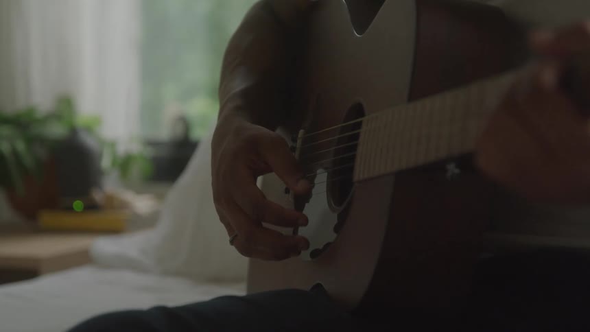 Based on the images from the video a person is seen playing an acoustic guitar the shot focuses on their hands strumming and picking the strings of the instrument the background is a slightly blurred indoor setting with a plant and natural light coming
