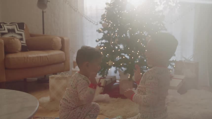 Siblings play in a living room both wearing pajamas with a christmas tree nearby