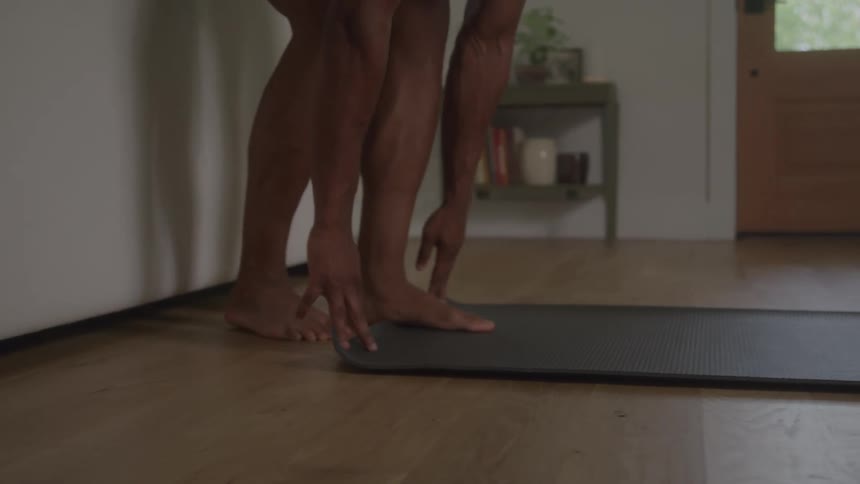 A man with a muscular build and a fade hairstyle wearing a white tank top and black athletic shorts enters a living space from the left unrolls a gray exercise mat onto the hardwood floor and then positions his body to begin a yoga exercise