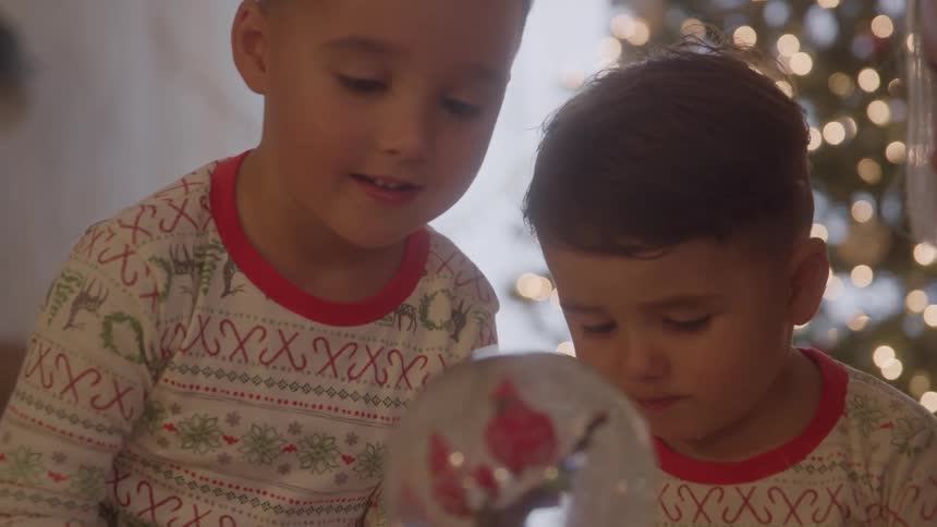 Two children wearing festive pajamas are seen looking intently at a snow globe with the older child pointing at the globe and the younger one looking at it with wide eyed excitement before both of them smile