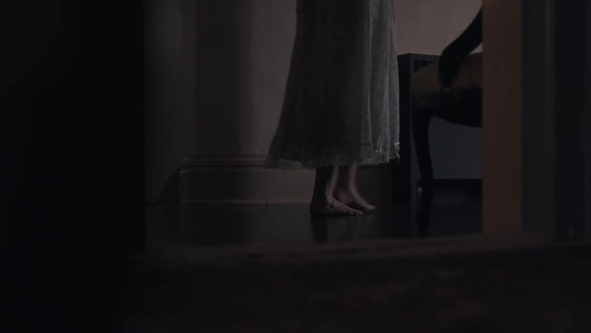 The video shows the feet of a person walking across a dark room the person is wearing a long lightcolored dress and is barefoot the room appears to be a bedroom or hallway with a chair visible in the corner