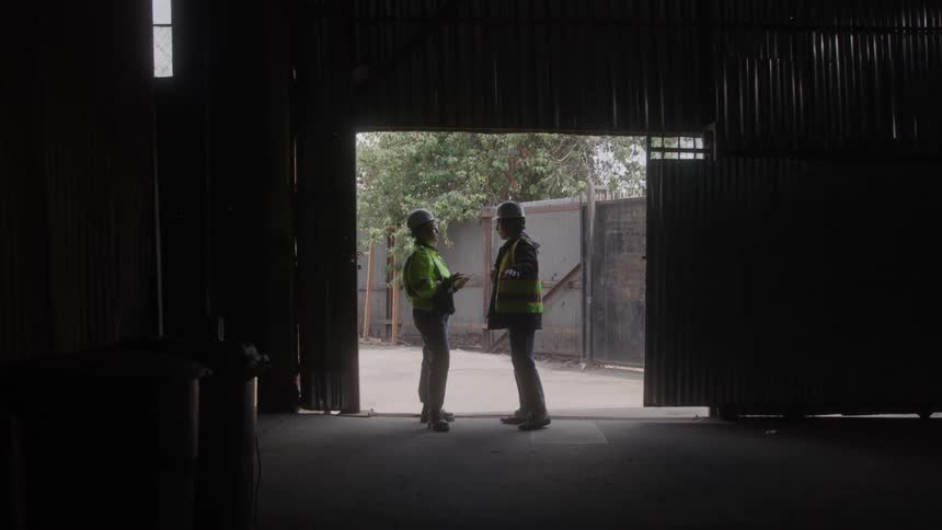 Two workers wearing helmets and reflective vests talking at the entrance of a warehouse