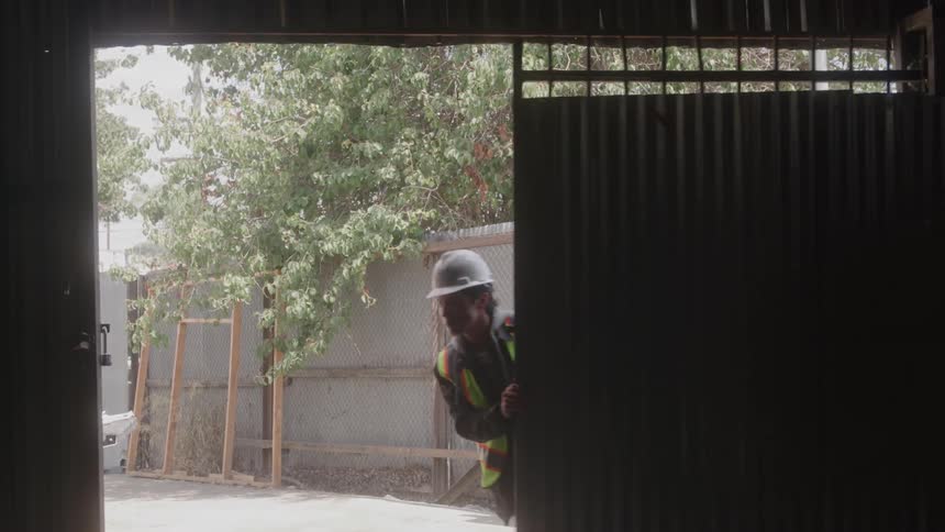 A worker wearing a hard hat and a highvisibility vest is seen from a distance the person walks from a brightly lit outdoor area into a dark indoor space closing a large metal sliding door behind them