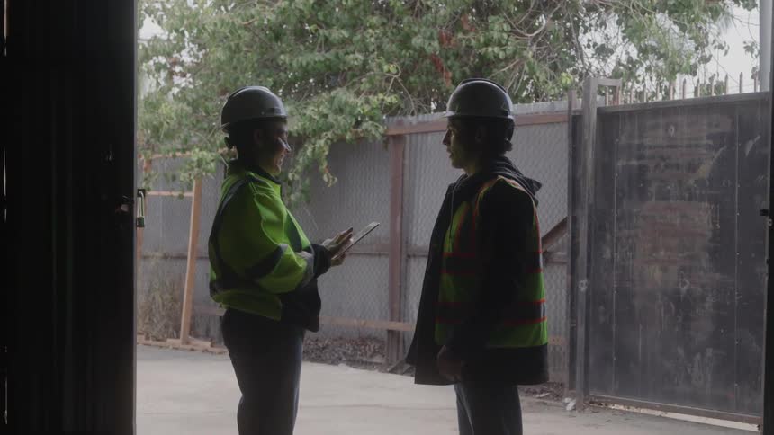 Two construction workers in full safety gear talk to each other on site then turn around to look behind them