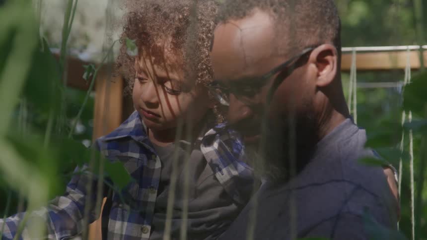 A person is shown from the chest up with a slight side profile they are holding a young child with curly brown hair in their arms both looking offcamera the person is wearing glasses and has a beard the scene appears to be outdoors in a garden or gr