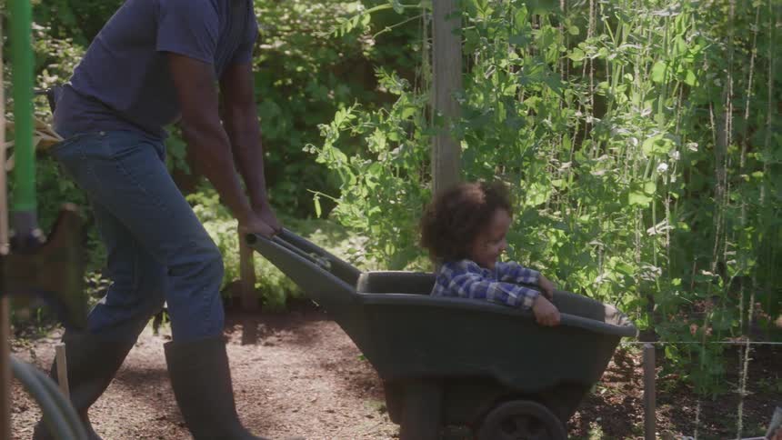 A man pushes a child sitting in a wheelbarrow down a path in a garden