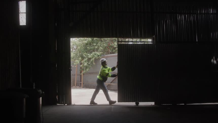 A long shot from inside a dark warehouse shows a person in a bright green highvisibility vest and a hard hat pushing a large metal door open the light from outside floods through the open door