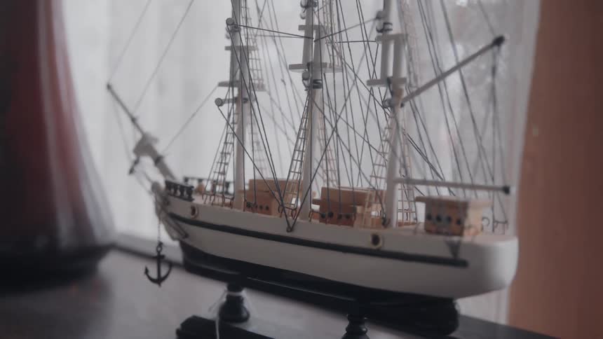 A replica of a sailboat sits on a table beside a window at midday