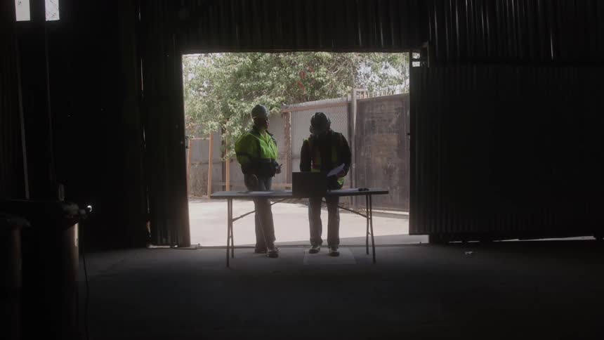 Two workers wearing helmets and reflective vests reviewing documents on a table