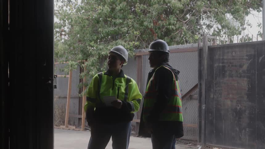 The video shows two construction workers standing outside wearing hard hats and highvisibility vests they appear to be discussing a project with one of them holding a tablet and pointing towards something offcamera