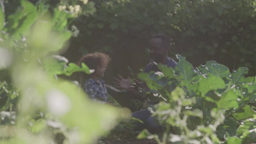 A blurry view of a man and a child with curly hair shows them both crouching down in a sunlit garden with green plants in the foreground