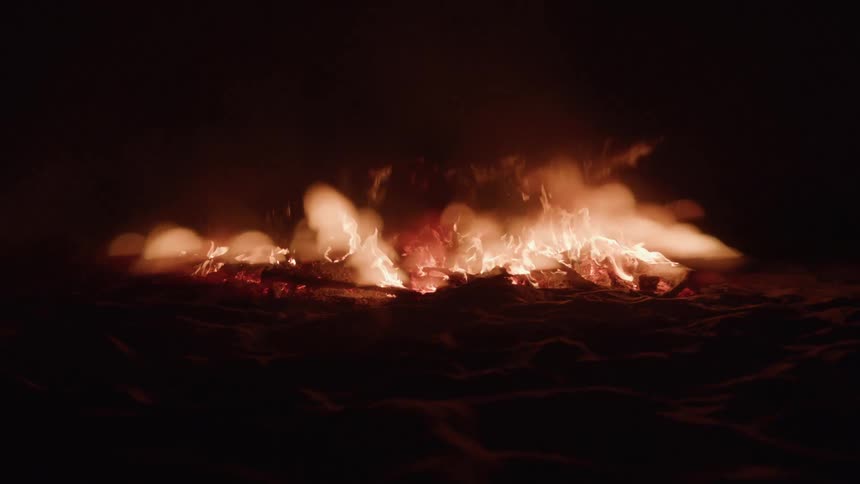 A bonfire burns brightly at night with red hot embers and orange yellow flames rising from a pile of wood and debris on a dark ground