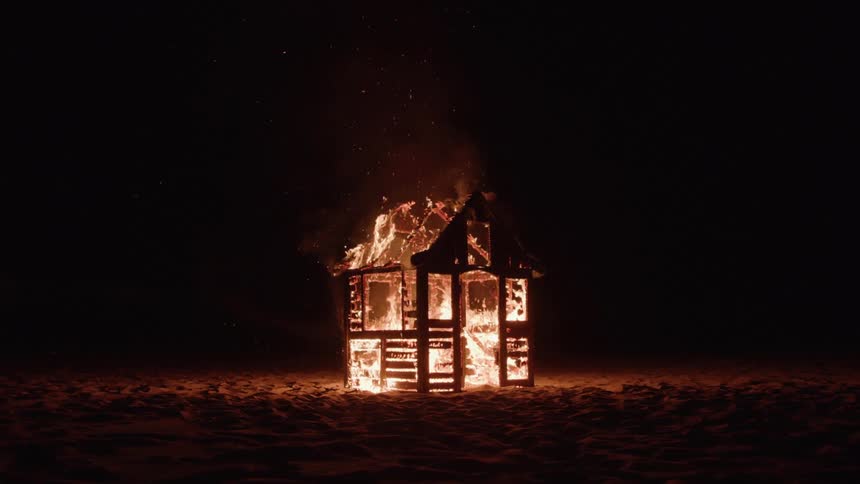 A small wooden structure burns intensely in the darkness flames illuminating the surrounding sand