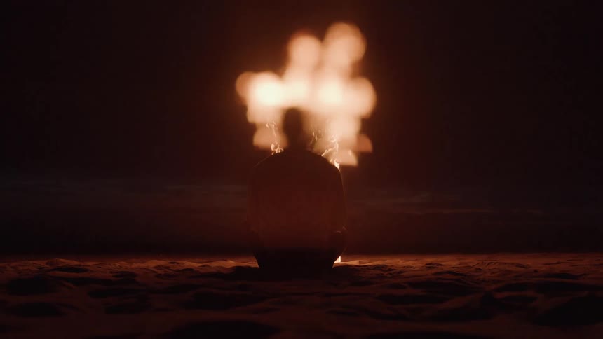 A person sits on sand at night with bright flames erupting behind their head casting an intense glow