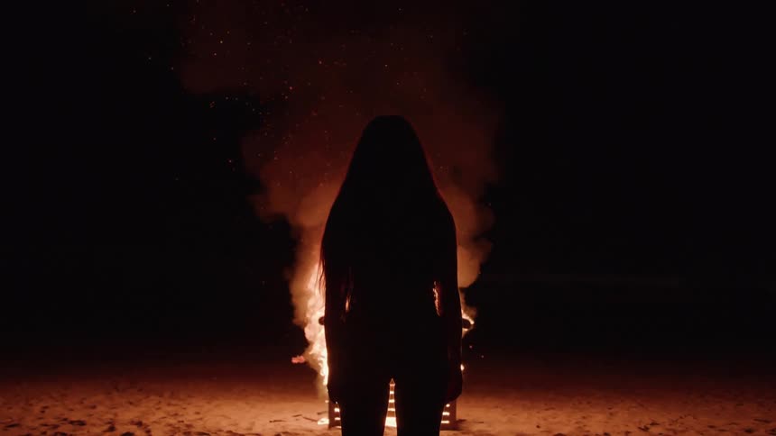 A persons silhouette standing in front of a bright bonfire at night with sparks and smoke rising