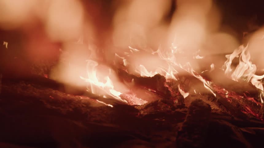 A close view of a large campfire shows wood logs burning with the red and orange flames glowing brightly