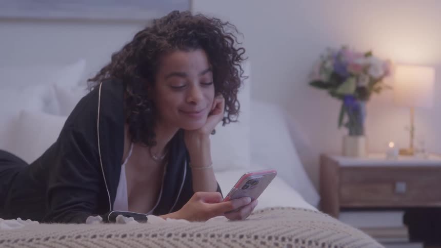 A young woman laying on a bed smiles while looking at her phone