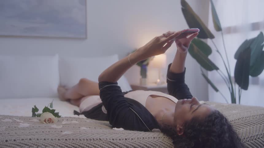 Woman lying on the bed looking at her phone with a rose beside her