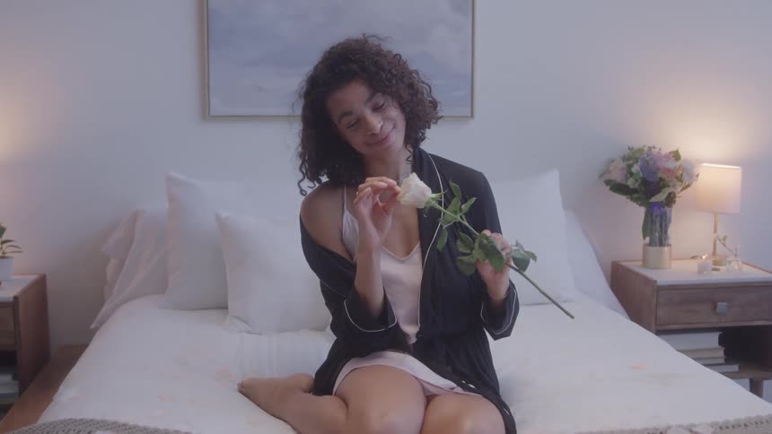 A smiling woman with dark curly hair is sitting on a bed in a bright modernlooking bedroom she holds a single white rose and looks at the camera while gently stroking its petals with her finger