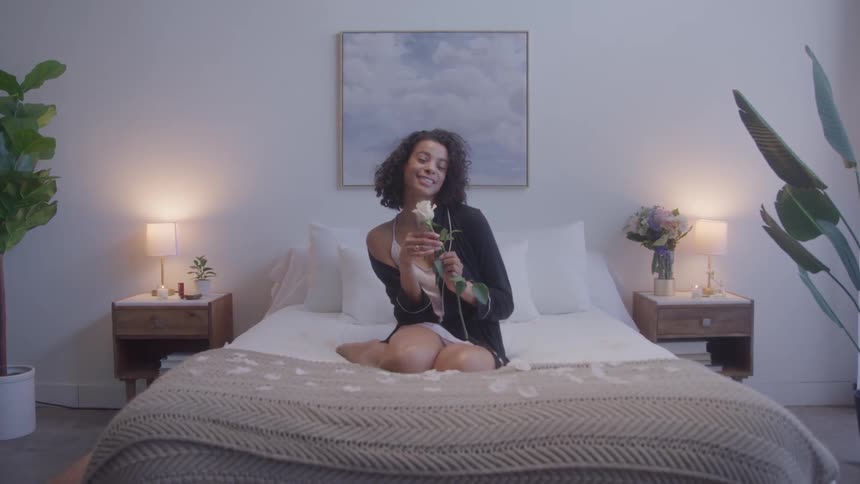 A young black woman perceives and adores a white rose flower while sitting in the middle of a bed in a bedroom