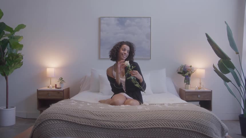 A young black woman adores a flower in her hands while sitting in the middle of a bed in a bedroom