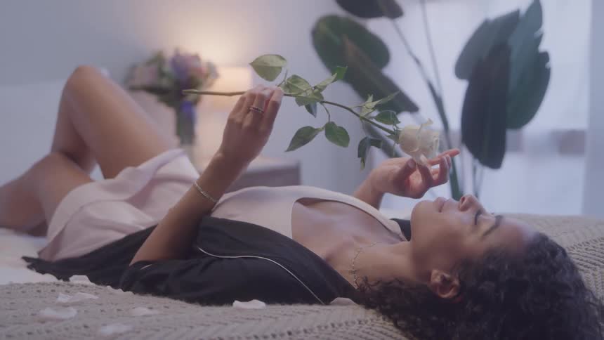 A young black woman admires and perceives a rose flower while lying down on a bed in a bedroom