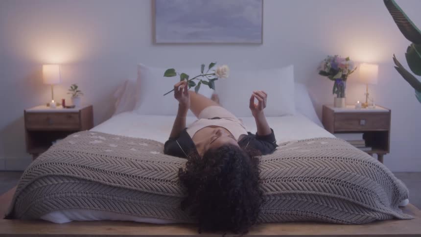 A young woman lies upside down on her bed in her bedroom facing the ceiling and holding a flower close to her nose as she smells it with lampshades on each side of the bed