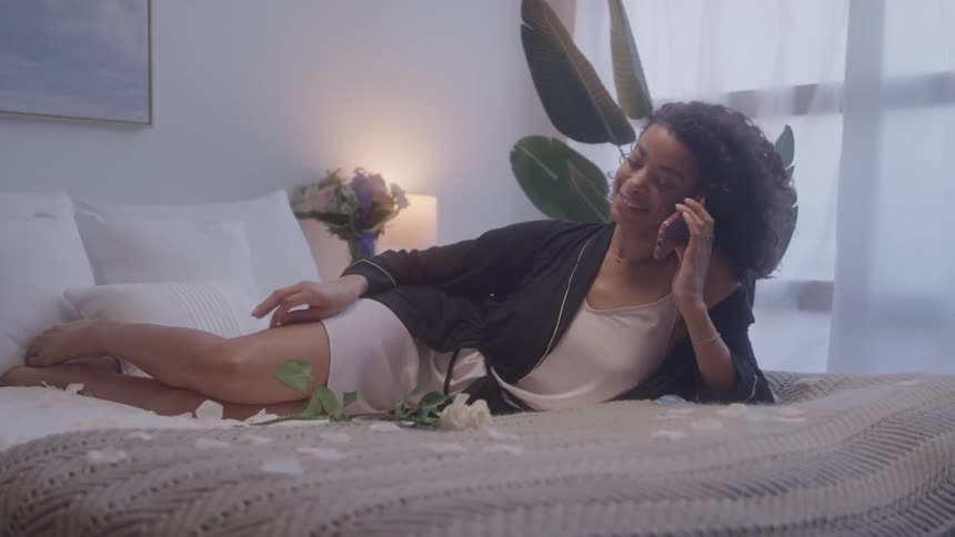 Young woman reclines on a bed strewn with flower petals while talking on the phone