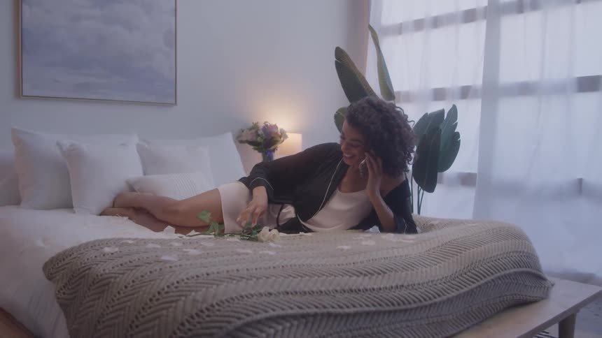 A woman with long curly dark hair lays on her side on a bed talking on a smartphone and holding a bouquet of flowers in one hand