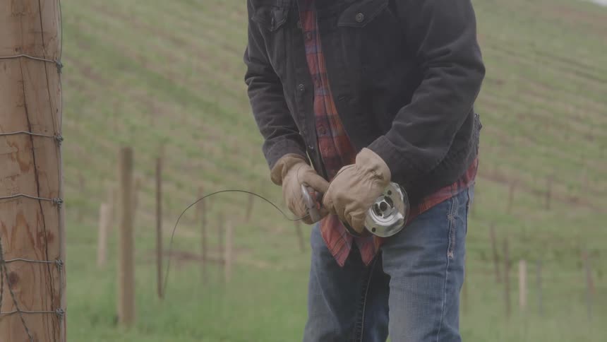 A person wearing a black jacket and a red and black flannel shirt blue jeans and brown gloves is seen from the waist up working on a wire fence