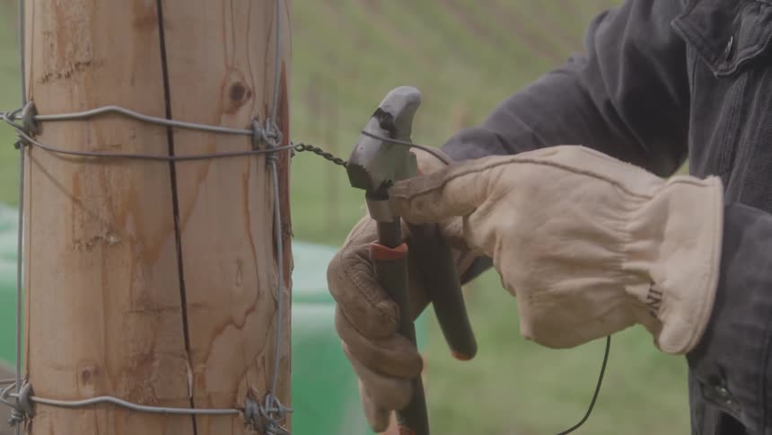 A closeup shot shows a persons gloved hands working on a fence post they are using a tool to tighten a section of wire that is wrapped around the wooden post