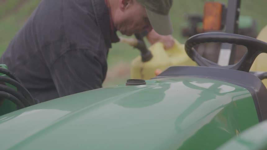 A person refuels a green tractor with a gas can