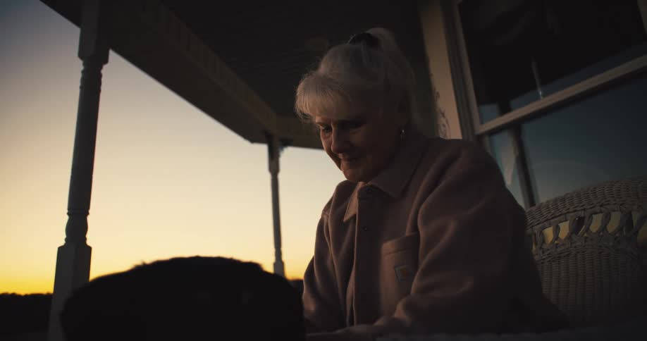 A woman with her white hair pulled up in a ponytail sits on a porch and looks out toward the golden sunset petting a dog in the foreground