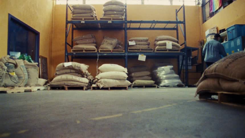 Two people are walking through a warehouse filled with large burlap sacks of grain and coffee beans stacked on the floor and on metal shelving