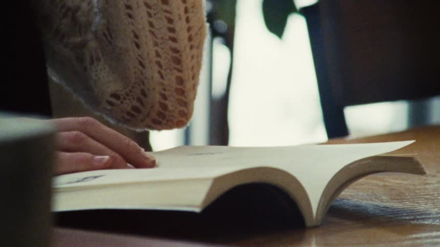 A woman sitting at a table while reading a book takes a drink from a coffee mug