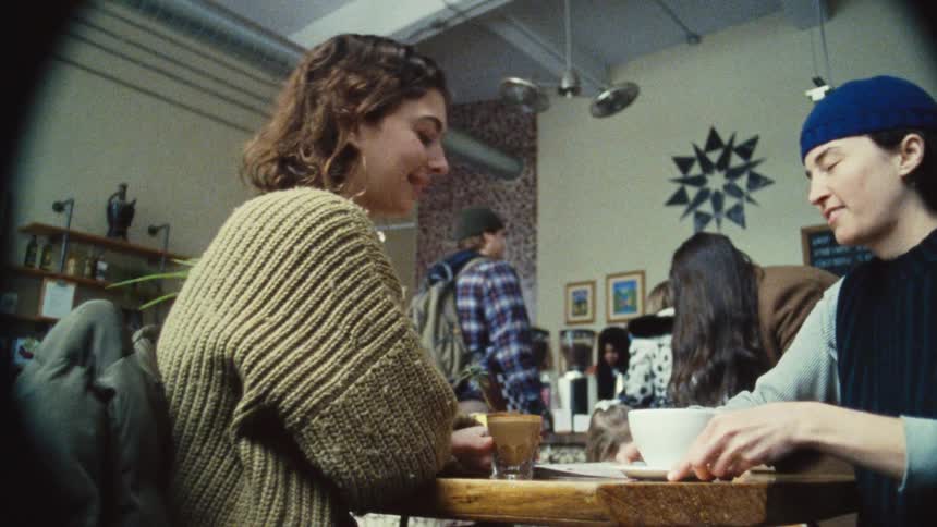 The video shows two people sitting at a table in a cafe looking at each other and smiling one person is wearing a chunky knit sweater and the other is wearing a blue beanie they are seen sharing a drink