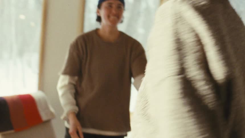 The video shows two people embracing and smiling happily in a room with large windows in the background they are wearing cozy sweaters and the person with shorter hair and a beanie is hugging the other person from behind