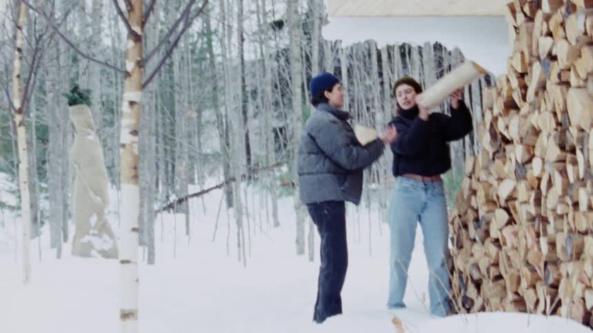 A woman picking up firewood and handing it to a man they are in the snow near a pile of logs