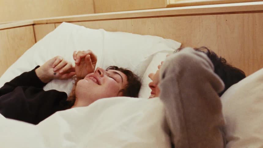 A couple in bed showing one woman slowly waking up with a smile as she looks at her partner