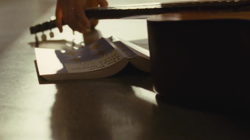 A person carefully places an acoustic guitar near a book on the floor